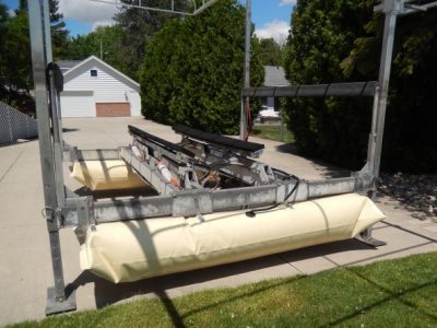 The Boat Lift Helper, featuring large beige air pontoons, sits on a driveway near trees and a white garage.