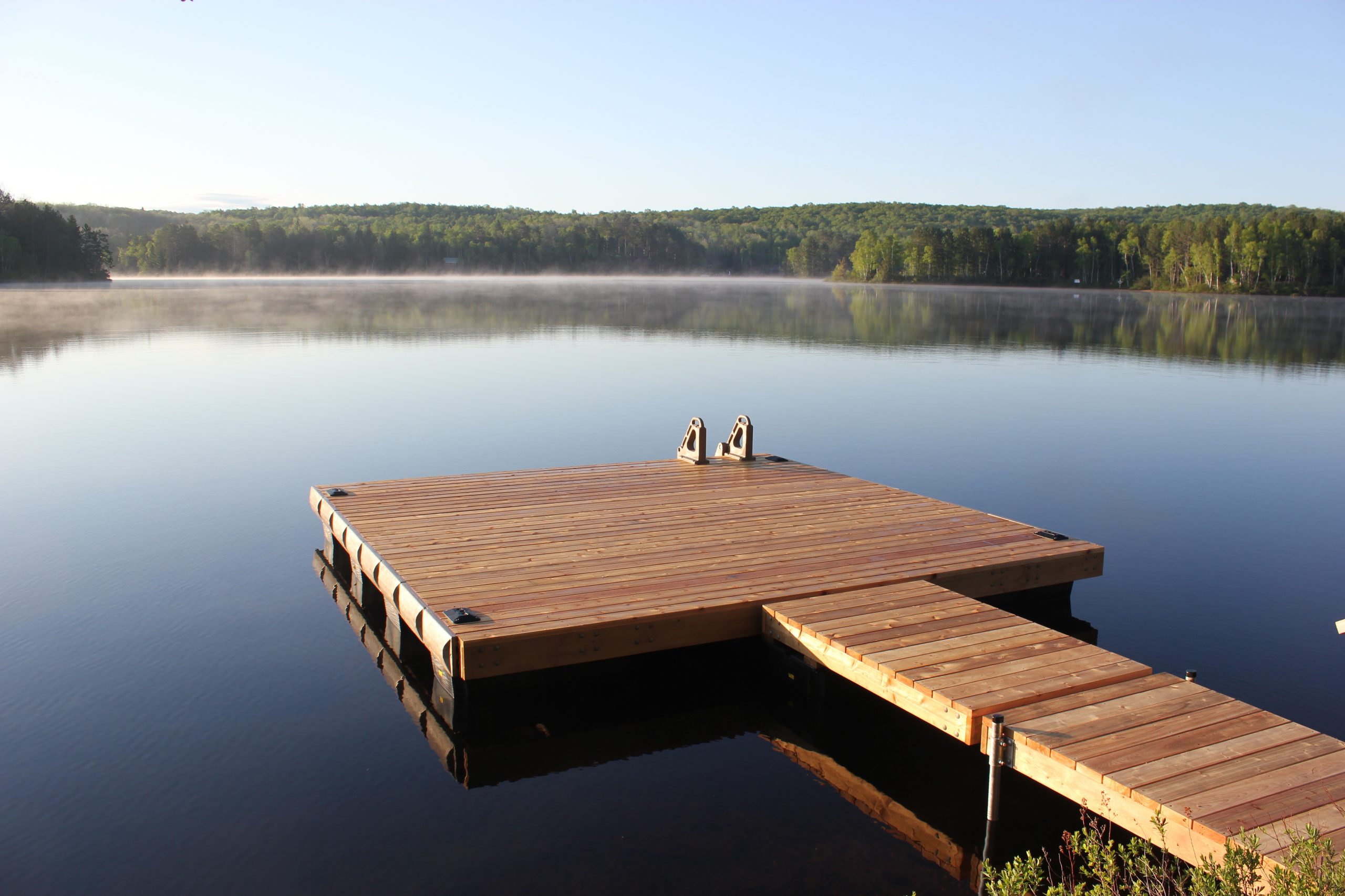16-x-16-AP-1 A wooden dock extends into a calm lake with mist rising and trees lining the shore in the background under a clear sky.
