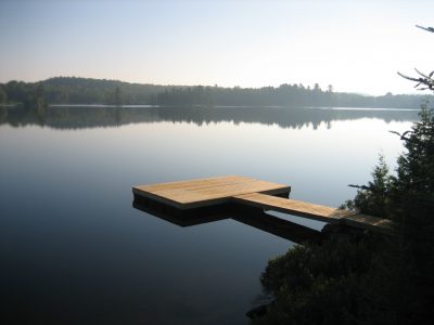 A wooden dock extends over a calm, reflective lake surrounded by trees under a clear sky.