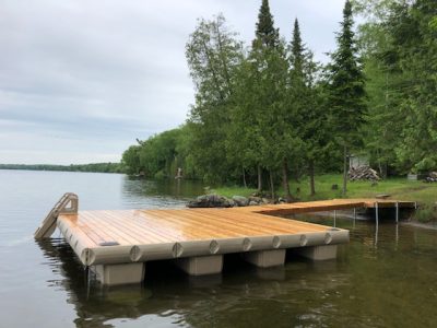 A wooden dock extends into a calm lake, surrounded by lush green trees and a rocky shoreline on a cloudy day.