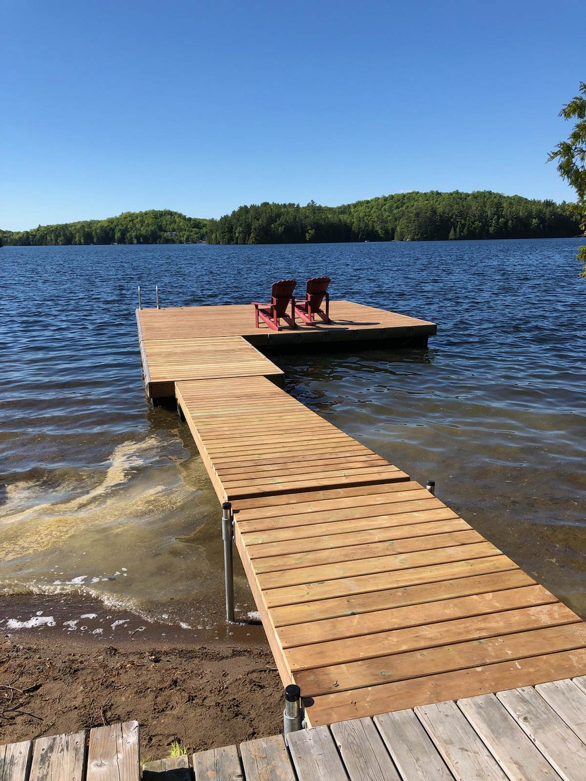 KNOW-YOUR-SWEET-SPOT A wooden dock extends over a lake with two red chairs at the end, surrounded by trees under a clear blue sky.
