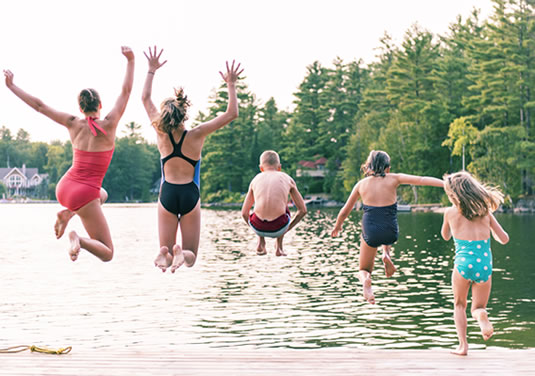 jumping Five children in swimsuits jump off a dock into a lake, surrounded by trees and lakeside houses on a sunny day.