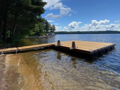 A wooden dock extends over clear lake water beneath a blue sky with clouds, next to a sandy shore and green trees.