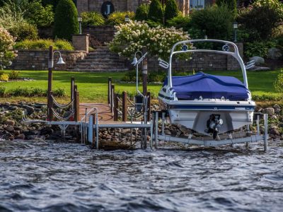 A covered speedboat is docked at a lakeside pier, with landscaped gardens and stairs leading up to a house in the background.
