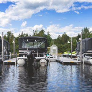 Three Boat Lift Canopies cover pontoon boats docked at a pier on a calm lake, surrounded by trees and a partly cloudy sky.