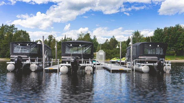 Three Boat Lift Canopies cover pontoon boats docked at a pier on a calm lake, surrounded by trees and a partly cloudy sky.