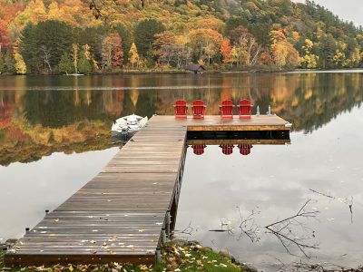 A wooden dock with red chairs extends over a calm lake, reflecting autumn trees with colorful foliage.